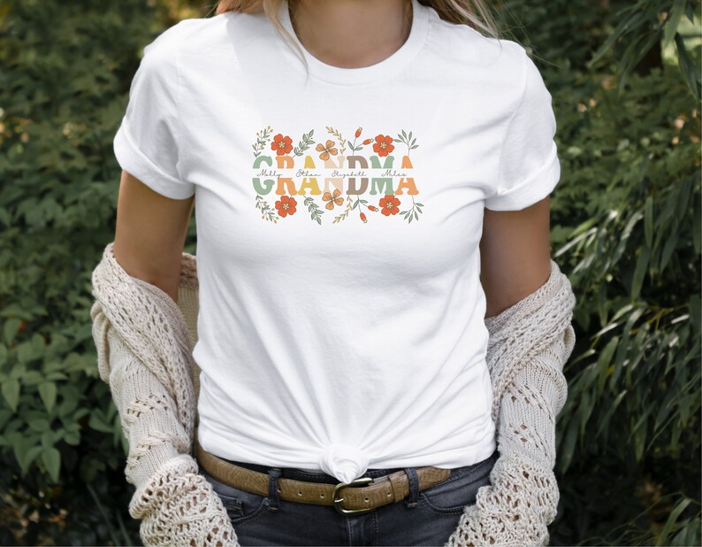 May include: White t-shirt with the word "GRANDMA" in colorful, retro-style lettering, surrounded by floral designs. Below the word "GRANDMA" are the names: Milly, Ethan, Elizabeth, and Mila. The shirt is a casual, everyday wear item.