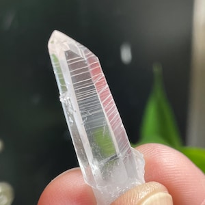 May include: A clear quartz crystal with a pointed tip and visible internal striations. The crystal is held between fingers, showcasing its translucent quality. The crystal's facets catch the light, highlighting its natural form.