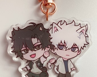 May include: A clear acrylic keychain featuring two cartoon characters. The character on the left is a black-haired cat wearing a white shirt and black trousers. The character on the right is a white-haired cat wearing a white shirt and black trousers. Both characters have large eyes and are smiling. The keychain is attached to a gold star-shaped metal ring.