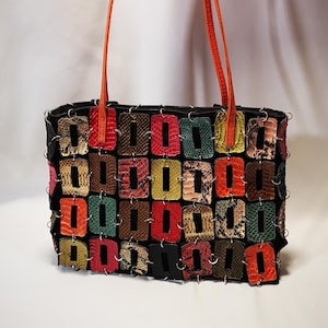 Peut inclure: Sac à main rectangulaire avec un design patchwork. Le sac présente une base noire avec des patchs rectangulaires colorés de différentes couleurs et motifs, dont le rouge, le vert et le marron. Il a des sangles orange.