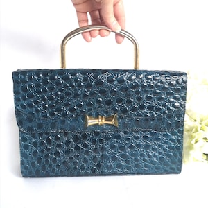 Puede incluir: Un bolso de mano de color azul verdoso con estampado de cocodrilo, con asa superior dorada y cierre de lazo dorado.