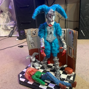 May include: A diorama featuring a blue-furred, jester-like figure with metallic silver limbs standing over a fallen doll in a green shirt and blue pants. A small gray rabbit figure sits nearby on a black and white checkered floor within a cardboard room.