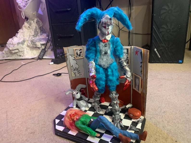 May include: A diorama featuring a blue-furred, jester-like figure with metallic silver limbs standing over a fallen doll in a green shirt and blue pants. A small gray rabbit figure sits nearby on a black and white checkered floor within a cardboard room.