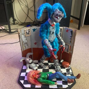 May include: A diorama featuring a blue, fuzzy, rabbit-like figure with metallic limbs and red accents standing on a black and white checkered floor. A smaller gray rabbit figure and a figure with pink hair in a green shirt lie on the floor.