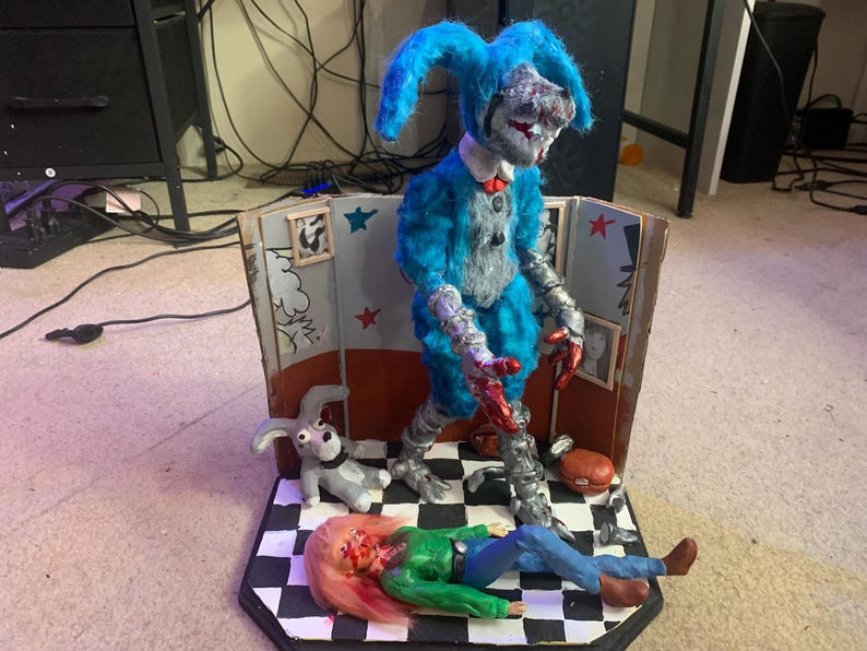 May include: A diorama featuring a blue, fuzzy, rabbit-like figure with metallic limbs and red accents standing on a black and white checkered floor. A smaller gray rabbit figure and a figure with pink hair in a green shirt lie on the floor.