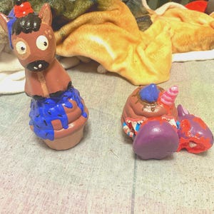 Op de afbeelding: Twee grillige keramische beeldjes. Eén is een bruin paardachtig wezen met een blauwe basis en een rode en zwarte hoed. De andere is een cupcake-vormig wezen met een paarse basis en een roze hoorn. Beide zijn handgeschilderd.