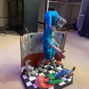 May include: A diorama featuring a blue, furry, wolf-like figure with metallic limbs standing on a black and white checkered floor. A small gray rabbit figurine and a fallen figure with pink hair and a green shirt lie on the floor near a cardboard wall.