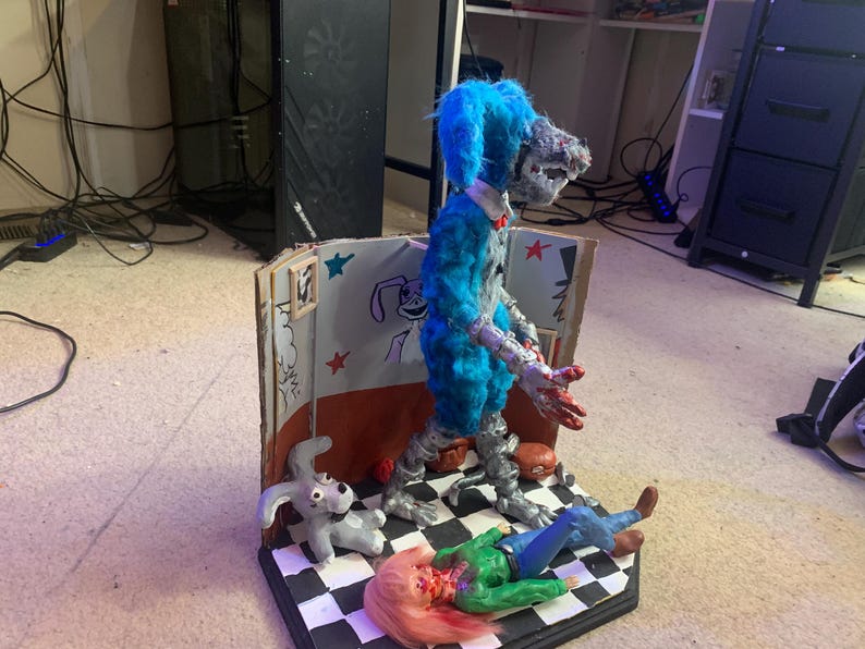 May include: A diorama featuring a blue, furry, wolf-like figure with metallic limbs standing on a black and white checkered floor. A small gray rabbit figurine and a fallen figure with pink hair and a green shirt lie on the floor near a cardboard wall.