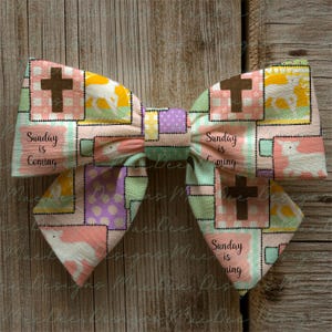 Może przedstawiać: Ręcznie robiona kokarda z patchworkowym wzorem. Zawiera krzyże, pastelowe kolory i napis "Sunday is Coming". Kokarda jest ozdobnym dodatkiem z centralnym węzłem.