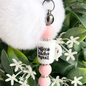 May include: White faux fur keychain with a pink and silver charm. The charm features a white coffee mug with the text "coffee teach repeat" and a small coffee cup icon.