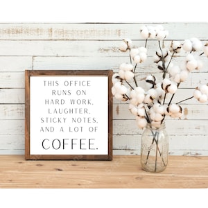 May include: A framed sign with a white background and black text that reads "This office runs on hard work, laughter, sticky notes, and a lot of coffee." The sign is sitting on a wooden surface with a vase of cotton flowers in the background.
