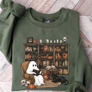 Puede incluir: Una sudadera verde con un gr&aacute;fico de un fantasma leyendo un libro en una silla junto a una estanter&iacute;a llena de libros. La estanter&iacute;a est&aacute; decorada con velas, calabazas y una calavera. Un gato negro est&aacute; sentado en el suelo junto a la silla.