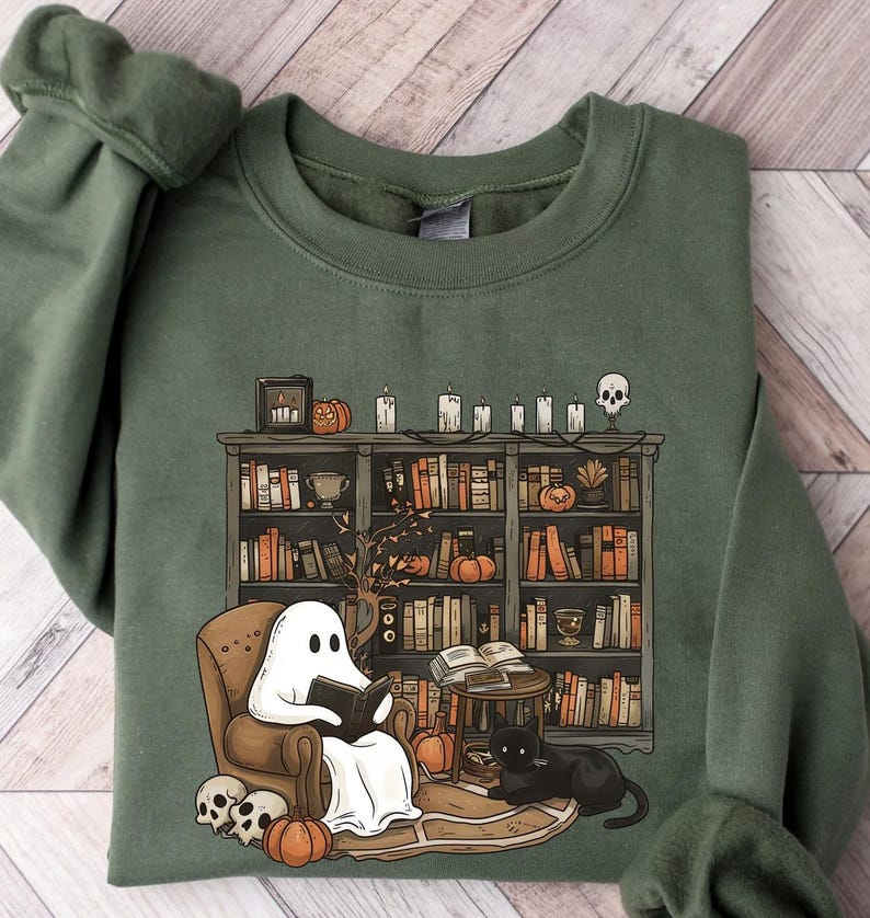 Puede incluir: Una sudadera verde con un gr&aacute;fico de un fantasma leyendo un libro en una silla junto a una estanter&iacute;a llena de libros. La estanter&iacute;a est&aacute; decorada con velas, calabazas y una calavera. Un gato negro est&aacute; sentado en el suelo junto a la silla.