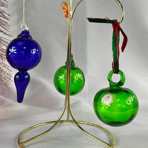 May include: Three glass Christmas ornaments on a gold metal stand. The ornaments are blue, green, and green. The blue ornament is teardrop shaped. The green ornaments are round.