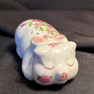 May include: A white ceramic pig figurine with pink accents and floral details. The pig is lying down with its eyes closed and has a pink bow on its head.
