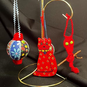 May include: A display of handmade ornaments. A red cat-shaped ornament with yellow floral accents and a blue bow tie hangs next to a round fabric ornament with a patchwork design. A red, woven ornament with green eyes is also displayed.