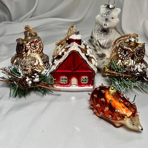 May include: A collection of glass Christmas ornaments in various shapes. Includes a red house with snow, a gray and white fox, two gold owls, and a brown hedgehog. Each ornament has a loop for hanging.