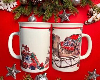 May include: Two white ceramic mugs with red trim. One mug features a Christmas scene with a sleigh and presents. The other mug has a blank white space. Both mugs have a red stripe around the top.