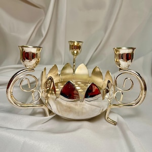 May include: A silver-colored candle holder with three candle cups and a central candle holder. The candle holder has a decorative, petal-shaped design around the base and scrollwork on the sides. The candle holder is made of metal.