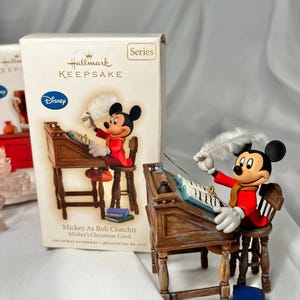 May include: A Hallmark Keepsake Christmas ornament featuring Mickey Mouse as Bob Cratchit. Mickey is seated at a wooden desk, wearing a red coat and yellow scarf, holding a quill. The ornament is approximately 3 inches tall.