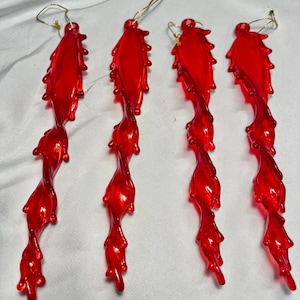 May include: Four red, icicle-shaped Christmas ornaments. Each ornament has a twisted, elongated design with a textured surface and a gold string for hanging. The ornaments are made of a translucent, glossy material.