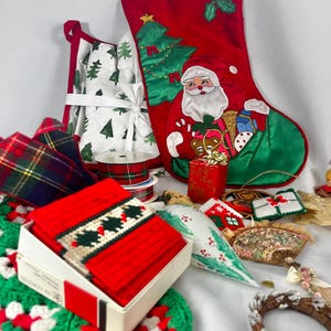 May include: A collection of Christmas decorations including a red stocking with a Santa Claus design, a white fabric stocking with a green tree pattern, red and green plaid ribbon, a white box with red and green crocheted squares, a white crocheted wreath, a white fabric cone with a red and green holly design, and a brown twig wreath with a red bow.