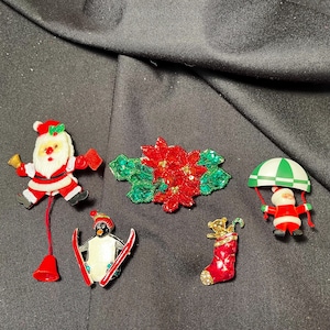 May include: Five Christmas-themed brooches, including a Santa Claus, a poinsettia, a penguin, a stocking, and a parachuting Santa Claus.