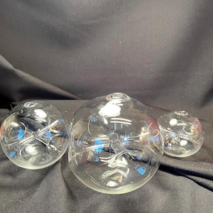 May include: Three clear glass spheres of varying sizes, each with a small opening at the top. The spheres are arranged in a row on a dark surface.