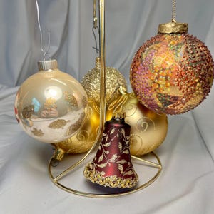 May include: A collection of Christmas ornaments in various shapes and colors, displayed on a gold-colored metal stand. The ornaments include a gold bell, a large textured ball with red and gold accents, and several gold and cream-colored spheres.