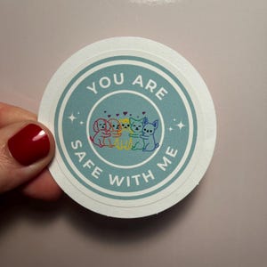 May include: A round sticker with a blue background and white border. The sticker features a circle with the text "You are safe with me" and a group of five cartoon dogs hugging each other. The dogs are rainbow colored.