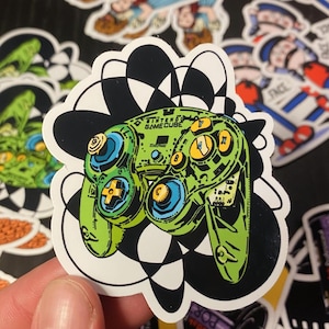May include: A green and black Nintendo GameCube controller sticker with a white background and a black and white swirl design. The controller has the text "Nintendo GameCube" on it.