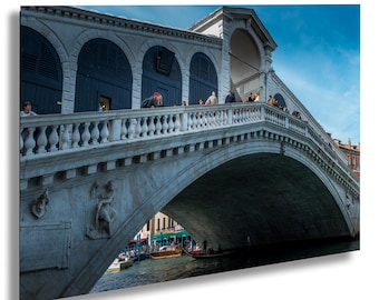 May include: A white stone bridge with an archway over a canal in Venice, Italy. People are walking across the bridge.