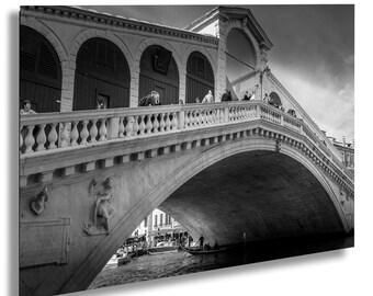 May include: A black and white photograph of a stone bridge in Venice, Italy. The bridge is arched and has a railing on either side. People are walking across the bridge. A gondola is in the water below the bridge.
