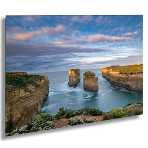 以下が含まれることがあります： 水からそびえ立つ3つの海食崖のある岩だらけの海岸線の全景。空はふわふわの白い雲のある鮮やかな青色。水は濃い青色で、岩は薄い茶色です。