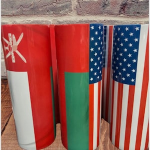 Op de afbeelding: Drie roestvrijstalen bekers met vlagontwerpen. De eerste beker heeft de vlag van Oman, de tweede de vlag van de Verenigde Arabische Emiraten en de derde de Amerikaanse vlag.