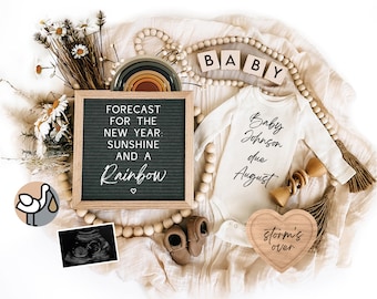 May include: A pregnancy announcement with a letter board that reads "FORECAST FOR THE NEW YEAR: SUNSHINE AND A RAINBOW". There is a white onesie with the text "Baby Johnson due August" and a wooden heart with the text "storm's over".