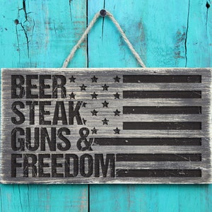 May include: A wooden sign with a distressed gray finish and black lettering. The sign features an American flag design with the words "Beer, Steak, Guns & Freedom".