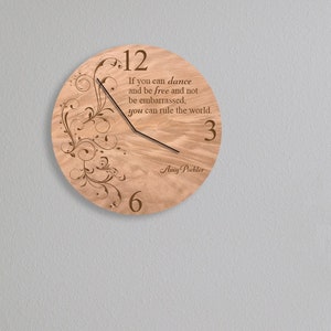 May include: A wooden clock with a quote about dancing and ruling the world. The clock has a light wood finish and a black hand. The quote is engraved on the clock face.
