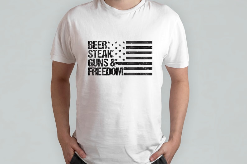 May include: White t-shirt with a black and white American flag design. The text "BEER, STEAK, GUNS & FREEDOM" is printed on the shirt.