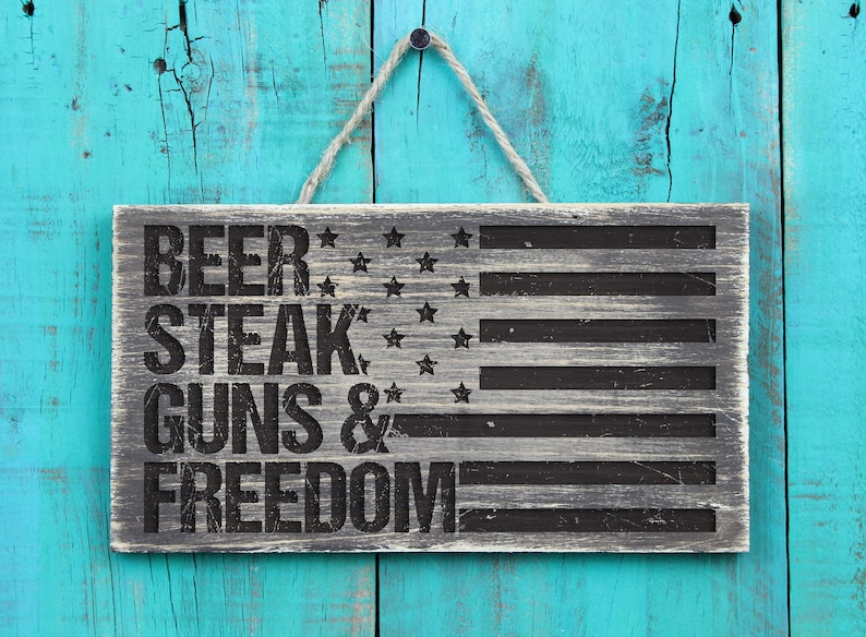 May include: A wooden sign with a distressed gray finish and black lettering. The sign features an American flag design with the words "Beer, Steak, Guns & Freedom".