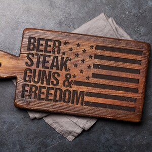 May include: Wooden cutting board with an American flag design and the text "Beer, Steak, Guns & Freedom".