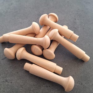 May include: A pile of unfinished wooden peg dolls, each with a rounded head and a cylindrical body. The dolls are made of light-coloured wood and are scattered on a grey surface. These peg dolls are suitable for crafting and DIY projects.
