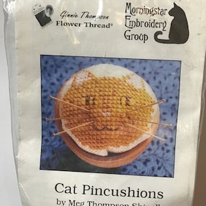 May include: A close-up of a cat-shaped pincushion with a yellow and white face. The pincushion is made of cross-stitch fabric and is sitting on a wooden base. The text on the packaging reads "Cat Pincushions by Meg Thompson Shinall".