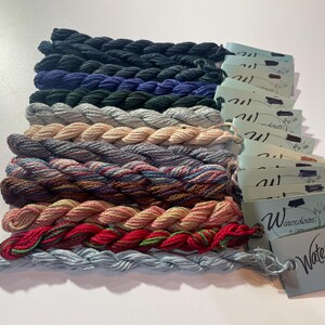 Könnte beinhalten: Eine Sammlung farbenfroher Stickgarne in verschiedenen Farbtönen, darunter Schwarz, Blau, Grün, Grau und Rot. Jedes Knäuel ist ordentlich angeordnet, mit Etiketten mit der Aufschrift "Waterlilies by Caron".