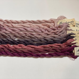 May include: A collection of six skeins of embroidery floss in a gradient of pink and purple hues. Each skein is tightly twisted, with a label reading "Alyce Schroth" and "Doylestown, PA 18901".