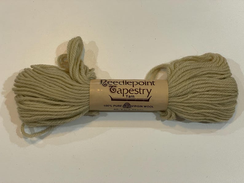 May include: A skein of beige Needlepoint Tapestry Yarn, made of 100% pure virgin wool. The yarn is wrapped around a tan cardboard tube with the brand name and product details printed on it. The yarn is a light, neutral color.