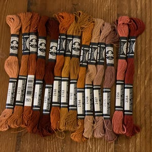 May include: A collection of DMC embroidery floss in various shades of orange, brown, and red. The spools are labeled with color codes and brand information. The threads are arranged on a wooden surface, ideal for craft projects.