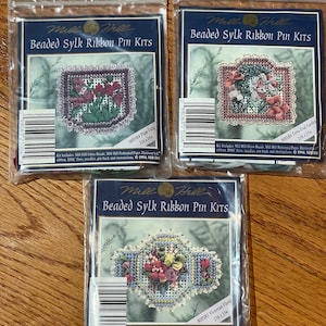 Può includere: Tre kit di spille per nastri di seta perlati Mill Hill. Ogni kit include perline di vetro, carta perforata, nastro di eredità, filo DMC, aghi, retrospina e istruzioni. I kit presentano motivi floreali in diversi colori e dimensioni. I kit sono etichettati con i nomi "Tulipano prugna", "Giardino di terrazza verde acqua" e "Floreale vittoriano".