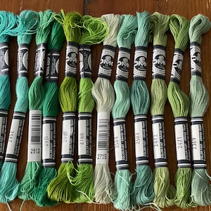 May include: A collection of green embroidery floss skeins in various shades, from teal to lime. Each skein is labeled with a color number and brand information. The floss is neatly arranged, showcasing a range of green hues.
