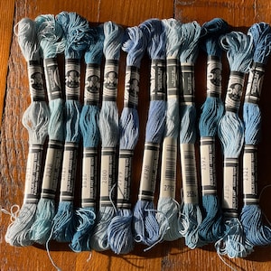 May include: A selection of blue embroidery threads in diverse tones, from pale to deep. Each spool is labelled with a number and brand details. The threads are arranged on a wooden surface, offering a visually pleasing arrangement for crafting projects.